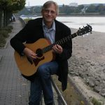 Christian Straube mit Gitarre und der der Beueler RHeinpromenade in Hintegrund