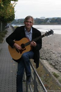 Christian Straube mit Gitarre und der der Beueler RHeinpromenade in Hintegrund