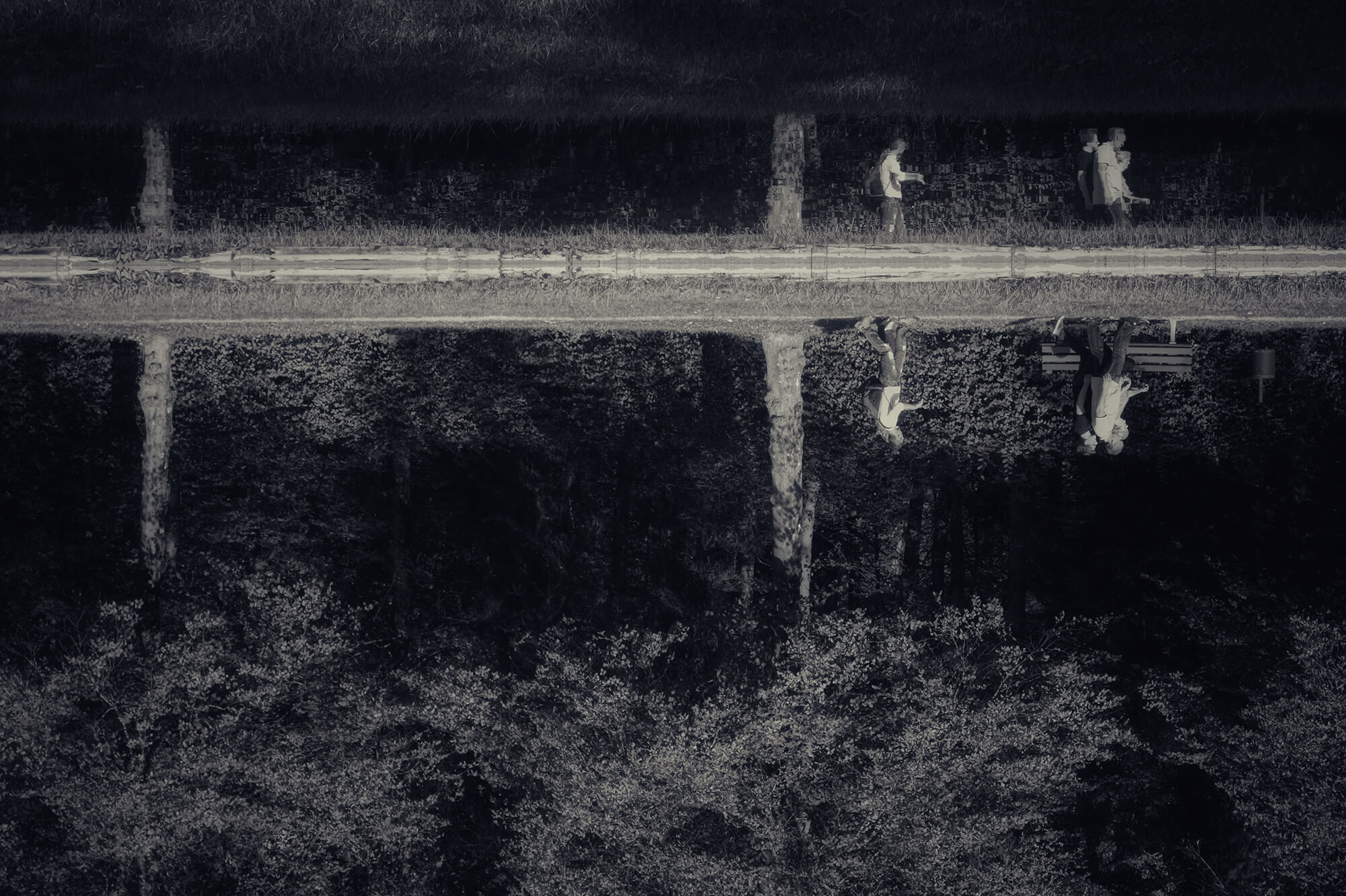 Schwarz-Weiß Foto über einen See hinweg, am anderen Ufer spiegeln sich Spaziergänger