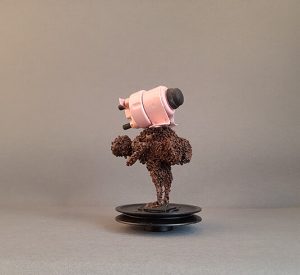 Crouching figure made of tiny metals scrapes, standing on a plastic socket balancing a pink big weight on its back