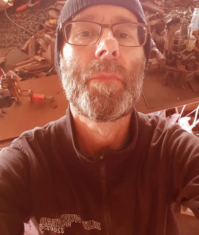 Selfie of Johan with beard and glasses with  the metal junk in the background that he makes his sculptures of
