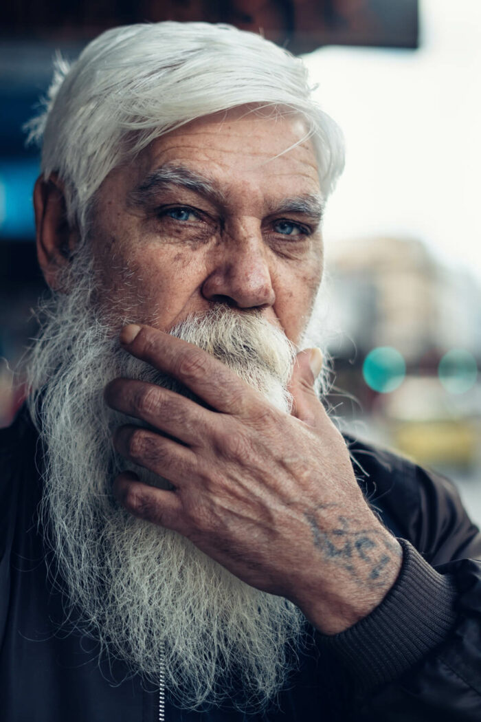 Old men with white hair and long white beard that he is scratching with a tatooed hand