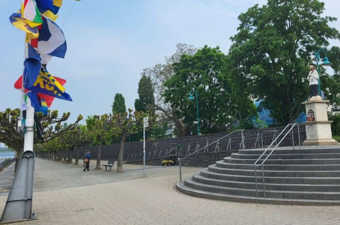 Blick von Norden mit Fahnenmast des Schiffervereins links, Statue des St. Nepomuk rechts und Denkmal Beueler Familie in der Mitte. Neugieriger Betrachter nähert sich ihr von links.