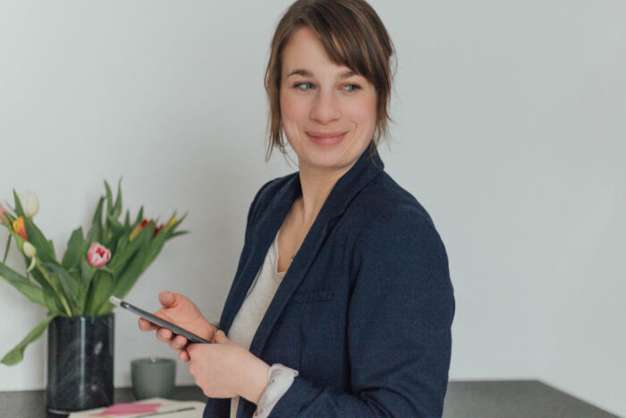 Anna Koschinski mit zusammengebundenen braunen Haaren im dunkelgrauen Blazer und weißem Shirt steht mit Smartphone in der Hand vor einem Tisch mit Blumenstrauß