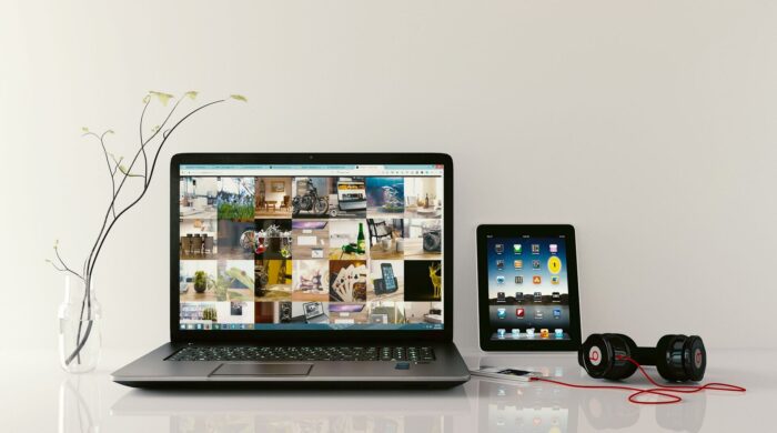 Ein geöffneter Laptop, ein Tablett und ein Kopfhörer stehen bzw. liegen auf einem weißen Schreibtisch vor einer weißen Wand, die mit einem Ast dekoriert ist