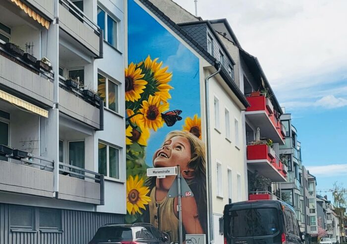 Haushohes Wandgemälde an einer vorstehenden seitlichen Häuserwand, das ein lachendes kleines Mädchen vor blauem Himmel zeigt, das sich an Sonnenblumen und einem Schmetterling freut