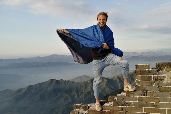 Oliver steht mit Schlafsack als Cape auf einer Ruine auf einem Berggipfel, im Hintergrund mehr Berge