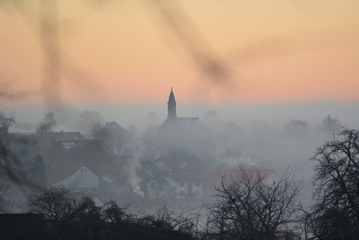 Blick bei Sonnenaufgang auf ein nebelverhangenes Dorf, aus dem der Kirchturm herausschaut