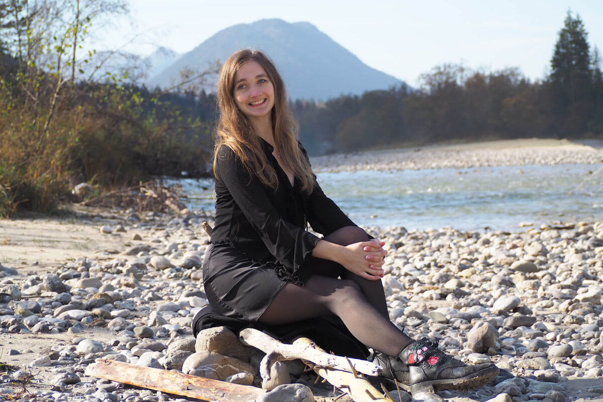 Lsa, eine junge Frau mit langen dunklen Haaren sitzt an einem Bach mit Bergen und Wald im Hintergrund