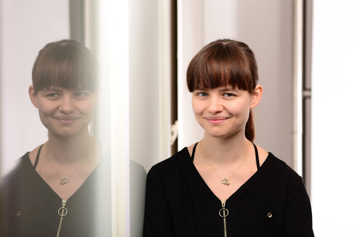 Edeline, eine junge Frau mit langen braunem Haaren spiegelt sich in einem Fenster
