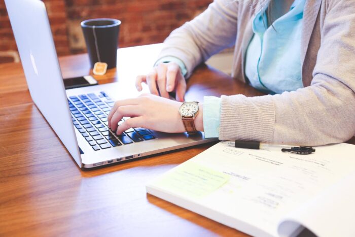Frauenhände auf Laptop, daneben Stfit und Papier und eine Tasse Tee