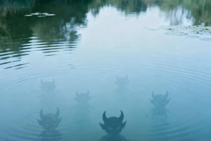 In einem See spiegeln sich der blaue Himmel und ein paar Baumkronen, unter der Wasseroberfläche sieht man die Silhouetten von kleinen Teufeln lauern