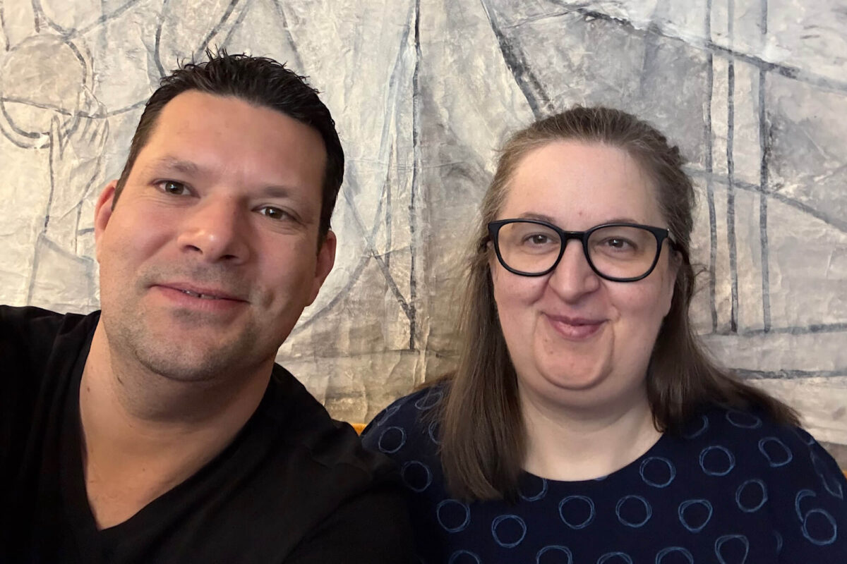 Ein kräftiger Mann mit kurzen braunen Haaren und eine Frau mit Brille und langen dunkelblonden Haaren lächeln in die Kamera