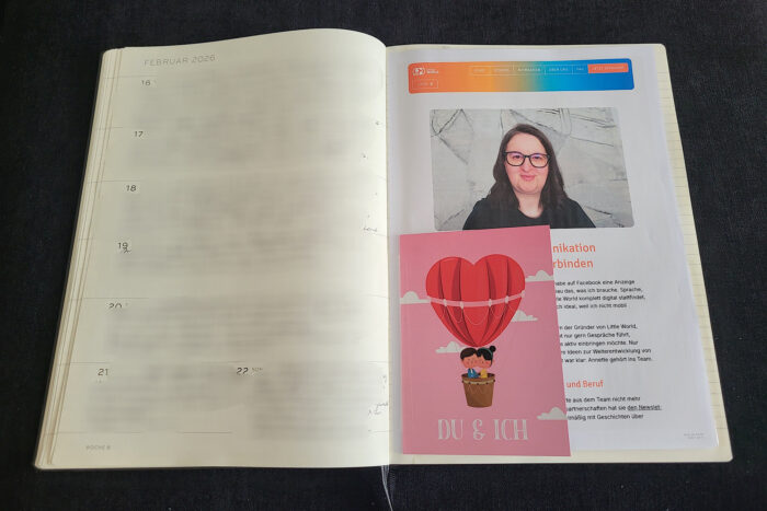 Aufgeklapptes Fotobuch mit verwischter Schrift links und eingeklebtem Ausdruck des Little World Artikels über Annette und einer Karte mit einem Liebesballon und der Aufschrift du und ich